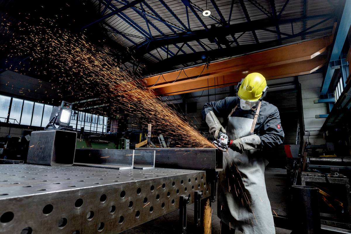 A worker in a hard hat is grinding metal, with sparks flying. He is wearing protective equipment in an industrial workshop.