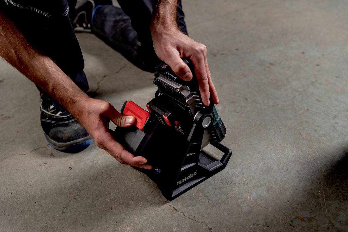 A person is changing the battery on a Metabo tool. The battery is being removed from a stand. The scene takes place on a concrete floor.