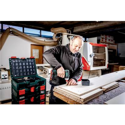 A man is working in a workshop with a drill on a piece of wood. In the background, there is a machine and toolboxes.