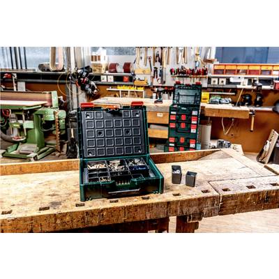 A workshop with wooden workbenches, tools hanging on the walls, an open box of screws and wall plugs on a table in the foreground.