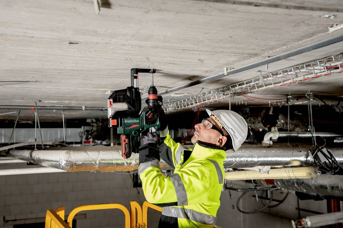 A worker in safety equipment is securing cables to the ceiling of an industrial building using an electric drill.