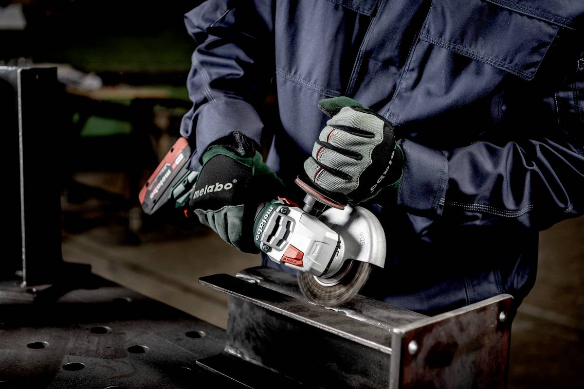 A person wearing protective equipment is grinding a metal piece with an angle grinder on a workbench in a workshop environment.