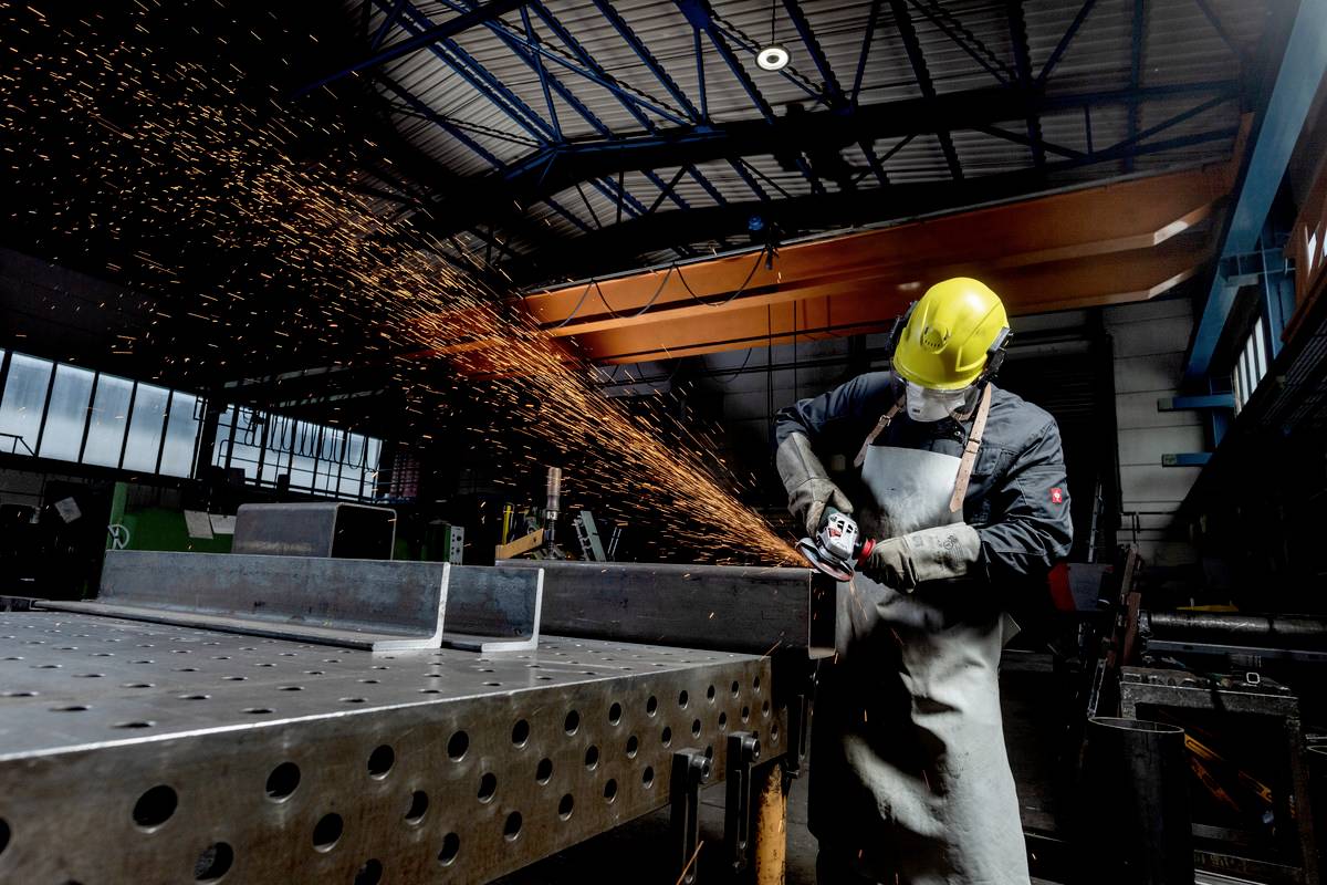 A worker wearing a hard hat and protective apron grinds metal, generating sparks in an industrial workshop environment.
