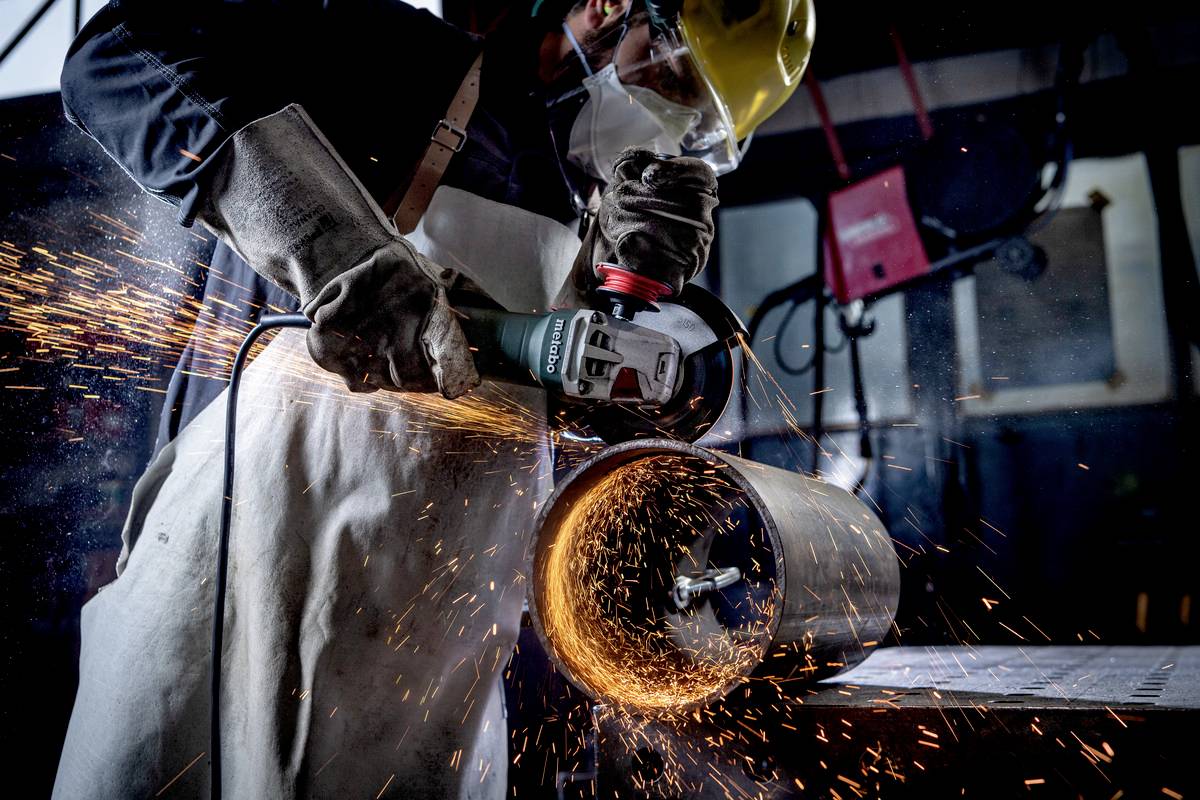 A worker wearing protective equipment grinds a metal pipe, sparks flying.