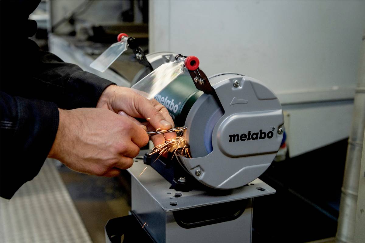A person is grinding a metal object on a metal grinding machine, with sparks flying. The machine part is labelled with "metabo".