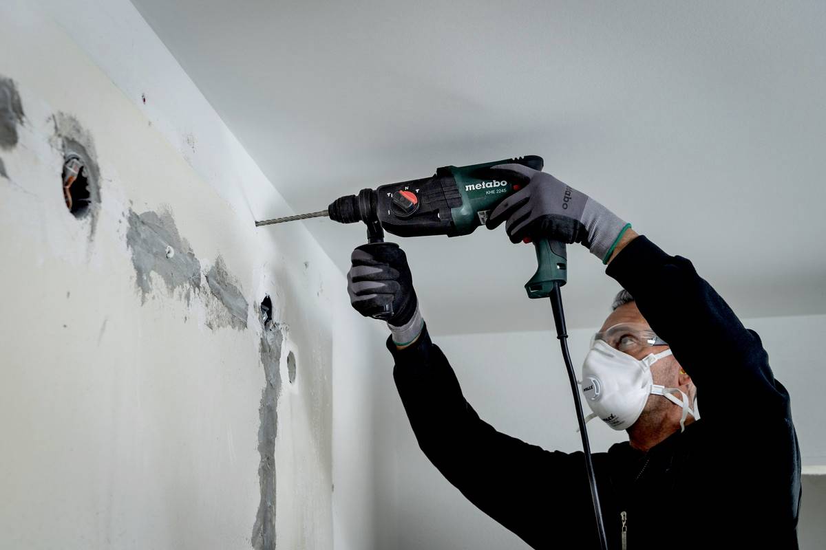 A man wearing a protective mask is drilling a hole in a wall with a drill, presumably during renovation work in a room.