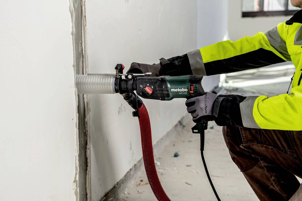 A person in work attire is using an electric tool with a suction hose to saw into a wall.