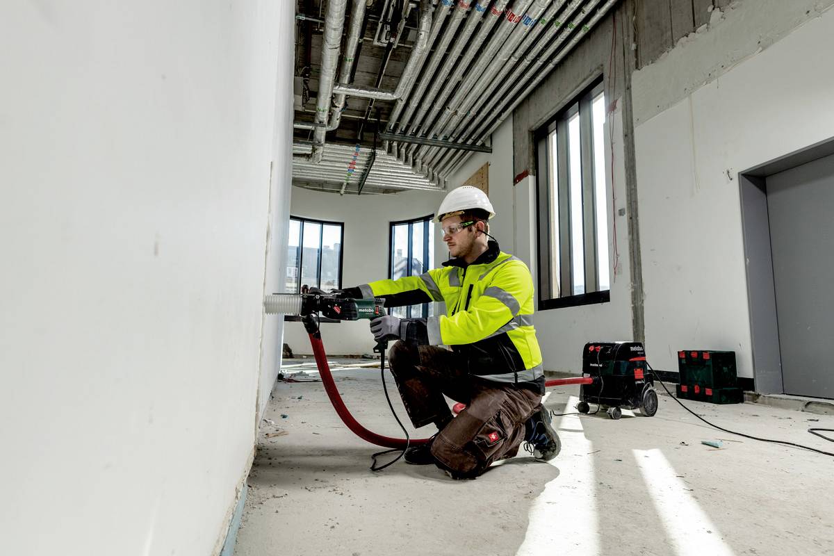 A construction worker in protective clothing is installing a cable in an unfinished building. Numerous pipes and conduits run above him.
