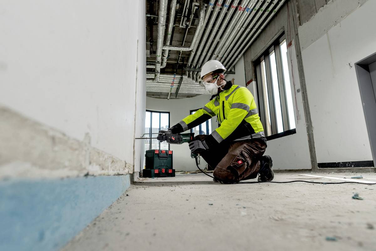 A construction worker wearing a hard hat and protective mask kneels in an unfinished room, operating a tool. Ceiling pipes and window frames are visible.