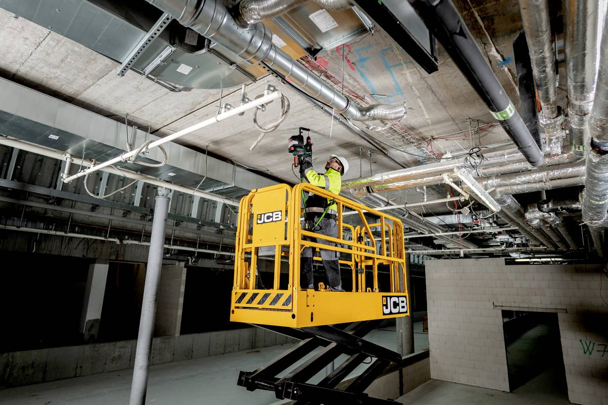 A worker in safety clothing is raising himself with a yellow hydraulic lift to the ceiling of an industrial building to carry out cable work.