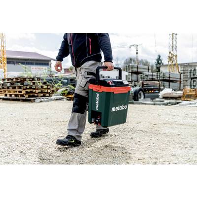 A man is carrying a green Metabo tool case on a construction site. The background shows building materials and cranes.