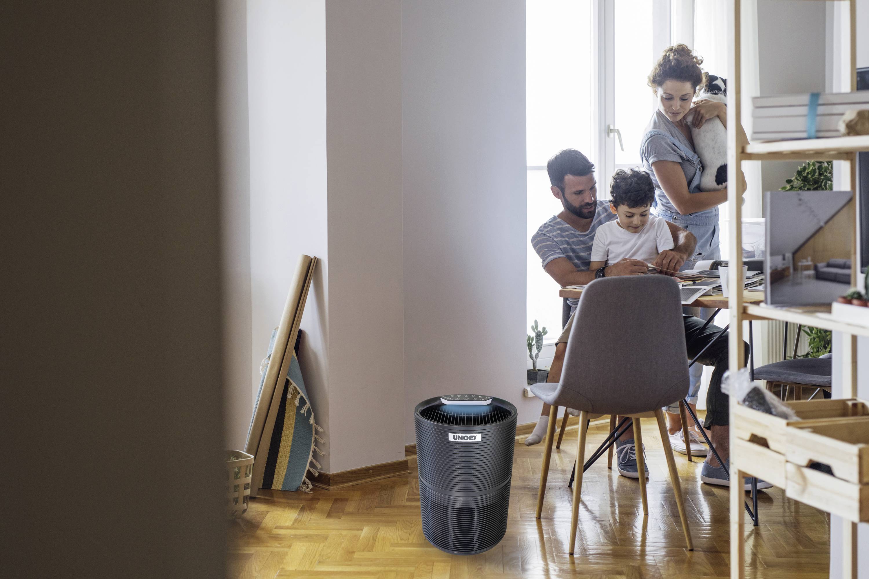 A family sits at a table with an air purifier in the foreground. In the background, a child is playing, while an adult holds a baby.
