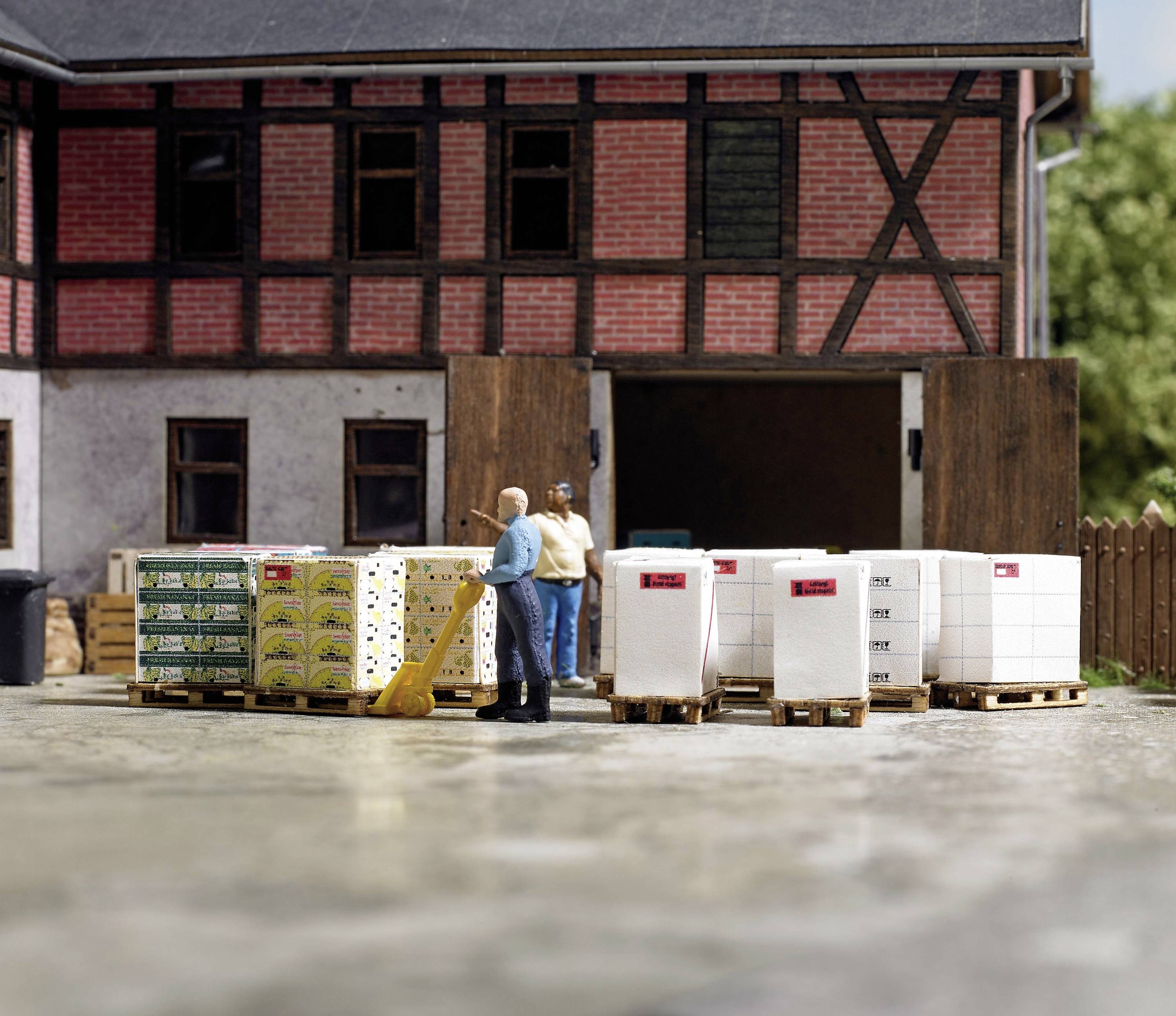 Two people are standing in front of a warehouse with stacked pallets. One person is holding a wheelbarrow. Buildings in the background.