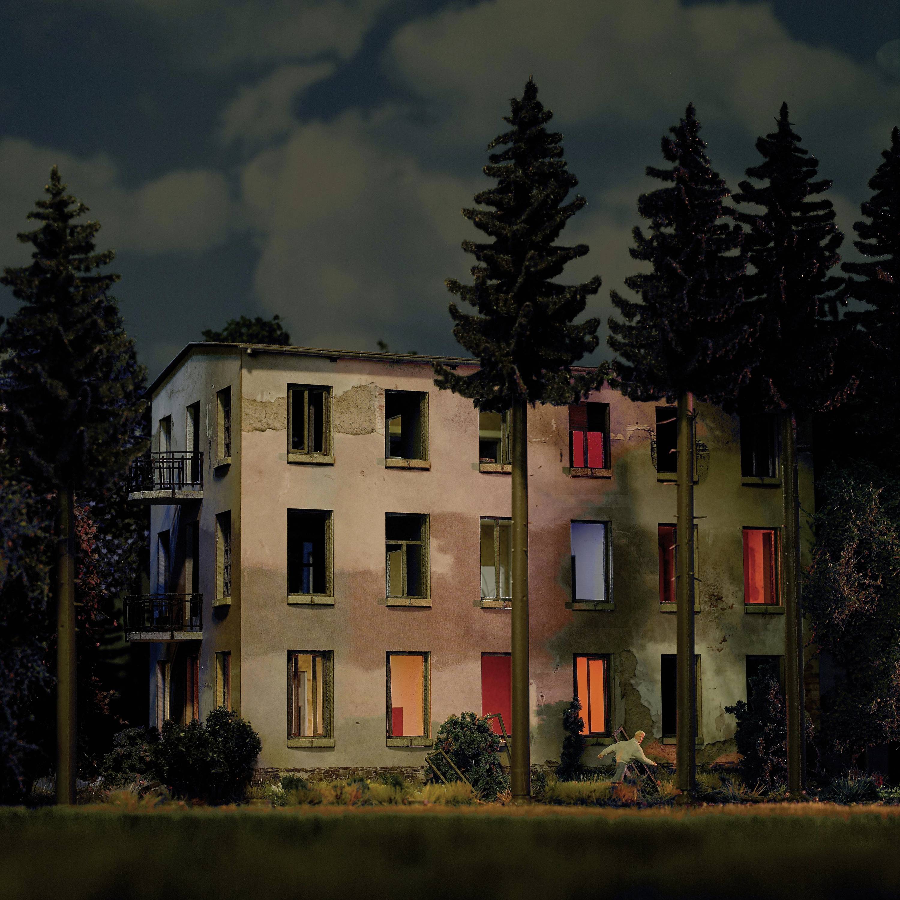 An abandoned building with broken windows is surrounded by tall trees. Light from within creates a mysterious impression.