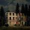 An abandoned building with broken windows is surrounded by tall trees. Light from within creates a mysterious impression.