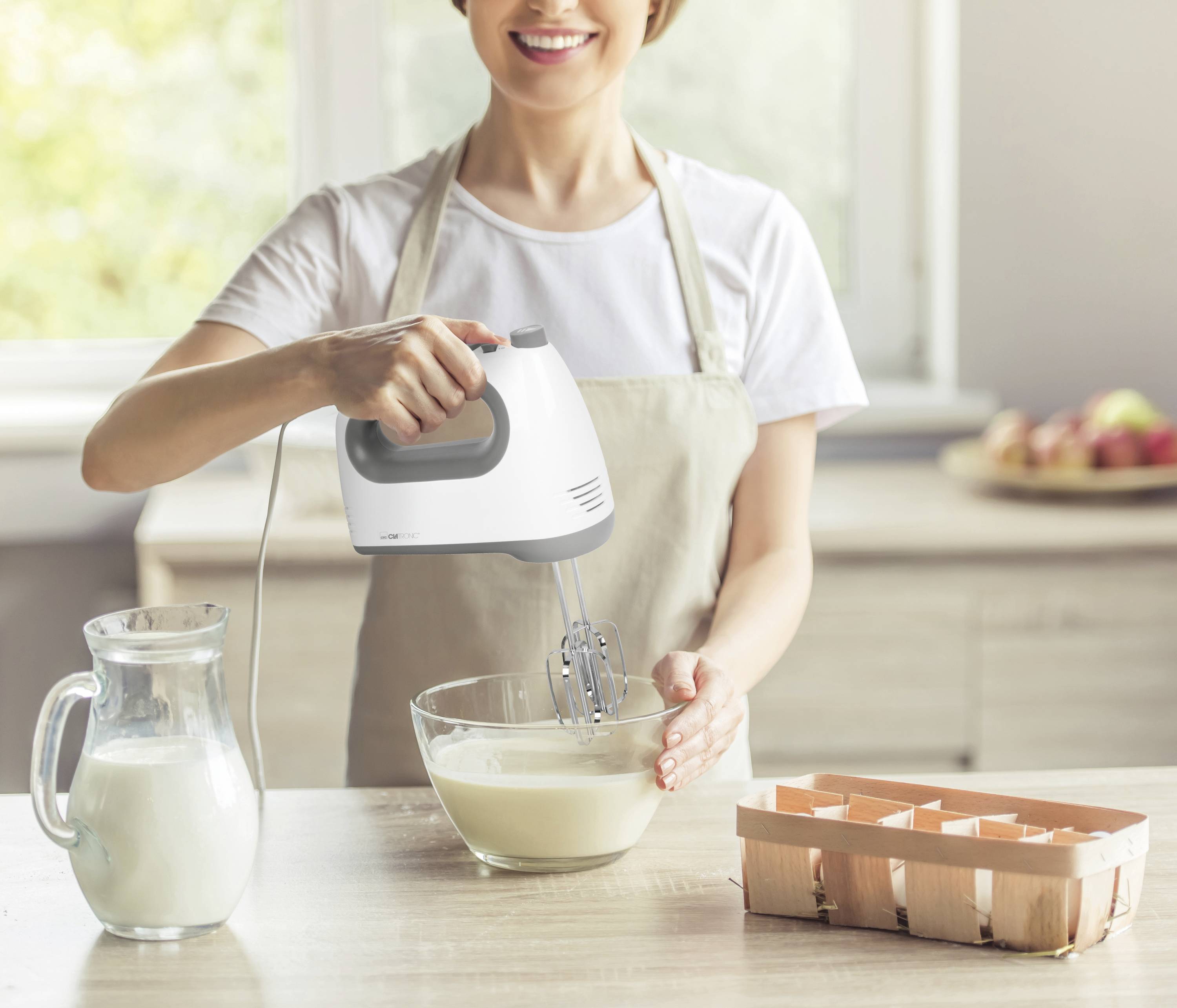 A woman is stirring dough in a bowl with an electric hand mixer in a kitchen. A milk jug and an egg carton are sitting on the table.