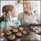 A girl and a woman are baking together. The girl is holding a tray of muffins, the woman is holding a baking tray with biscuits. They are laughing.