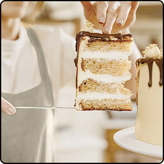 A person is cutting a slice of multi-layered cake with chocolate ganache. The setting is a bright, cosy kitchen.