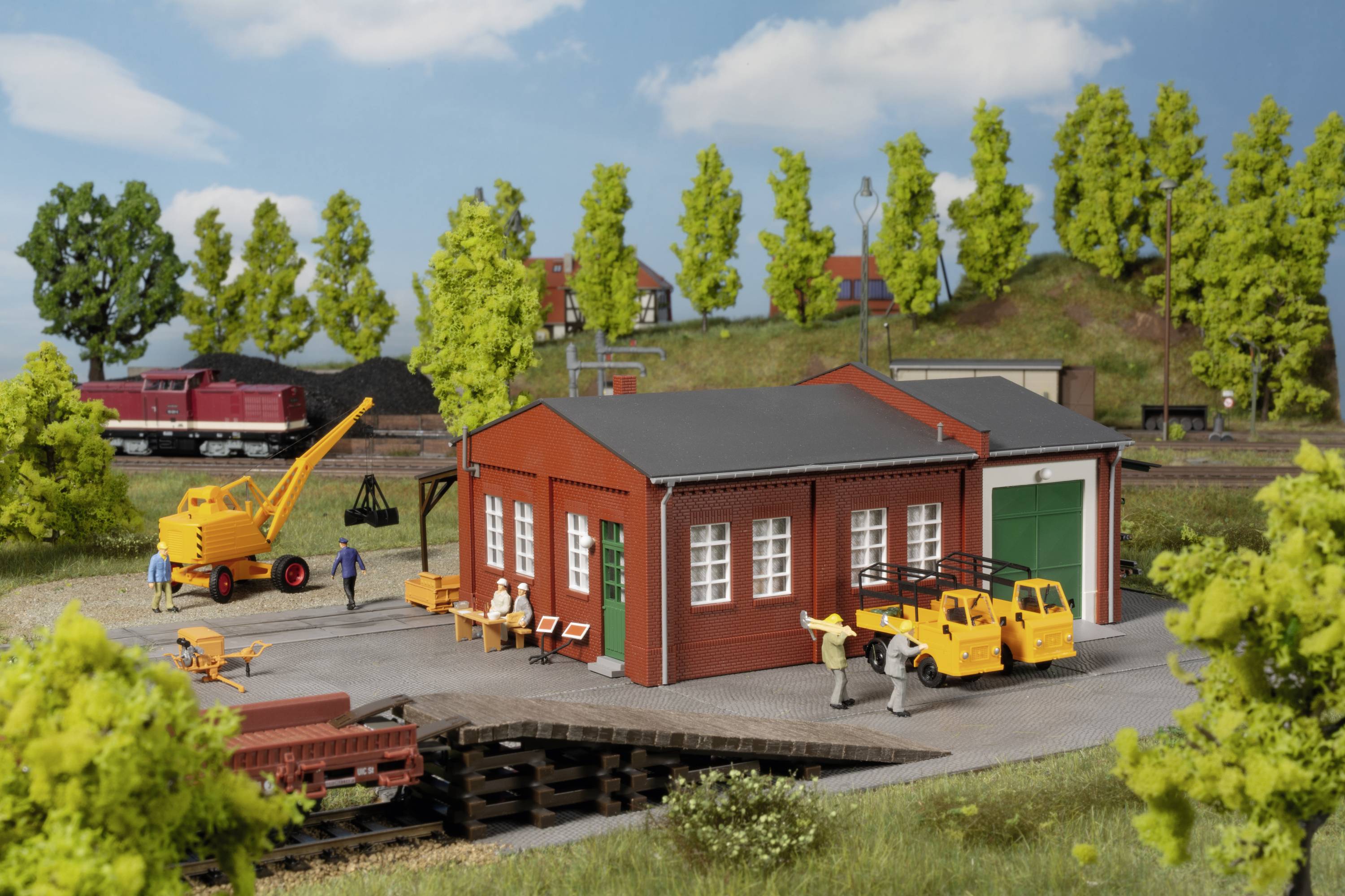 Small model construction scene with a red building, yellow construction machinery and people. Trees and a train are visible in the background.
