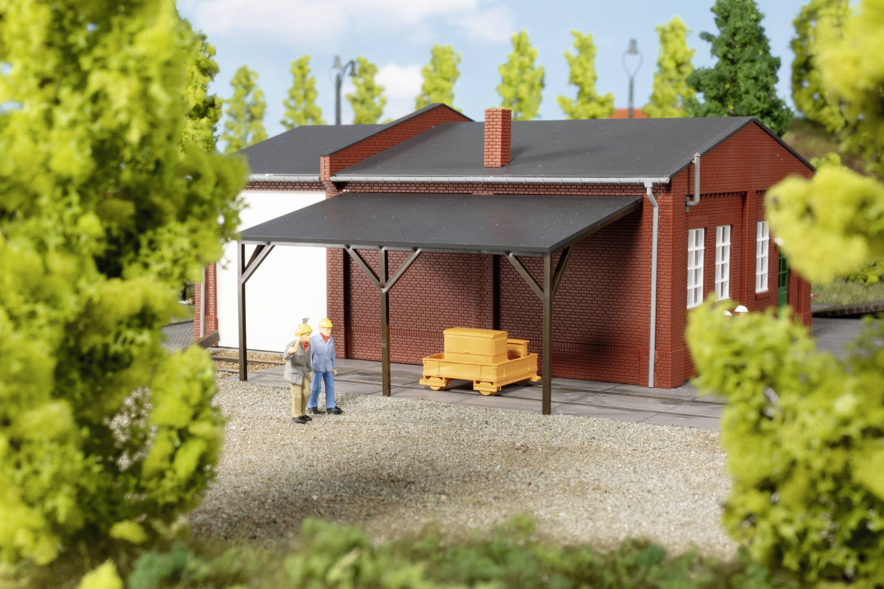 A model railway building with a covered area, two people and a small goods van in the foreground, surrounded by trees.