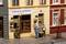 Three figures stand in front of a tobacco and whisky shop. One man is carrying a crate, while another stands beside him. A sign reads 'Tobacco & Whisky'.