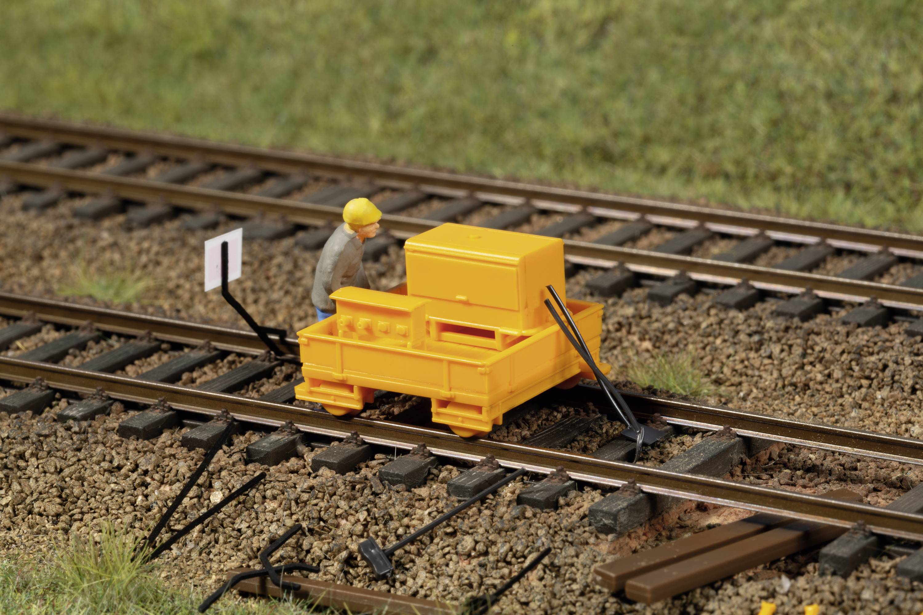 A yellow miniature vehicle on a model railway track, controlled by a figure wearing a yellow hard hat, demonstrates track maintenance.