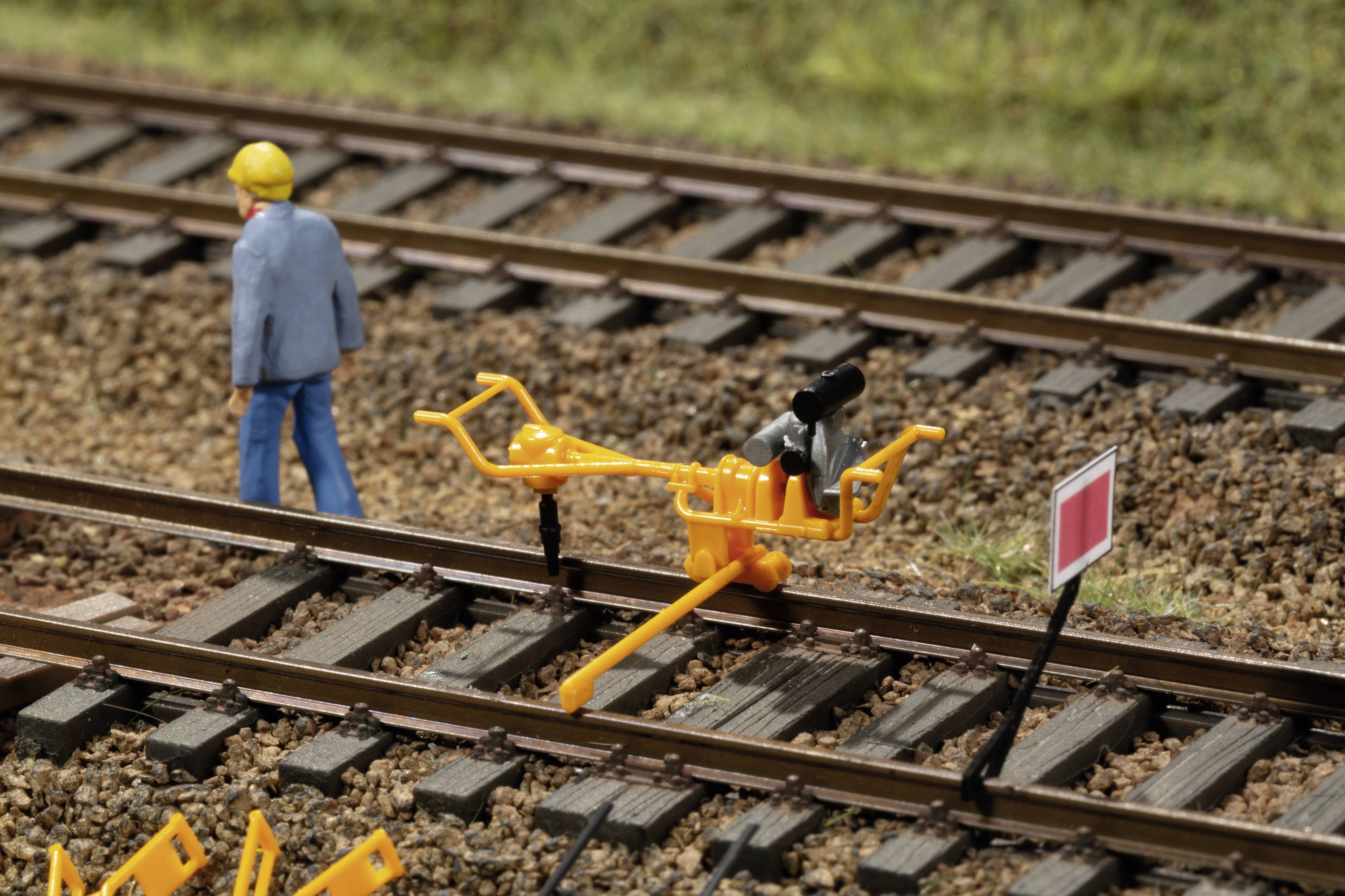 A miniature figurine of a construction worker stands beside a yellow rail device on a model railway track. A small sign is visible.