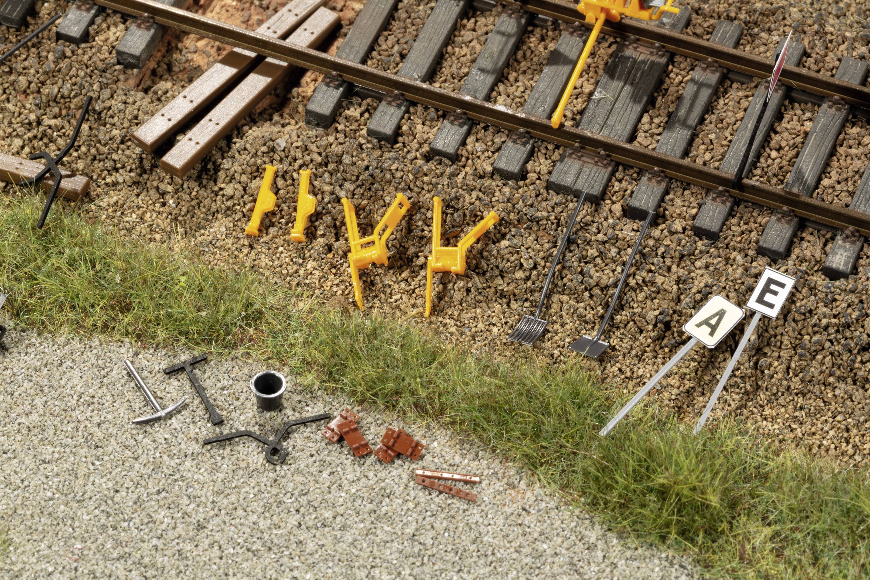 Model railway scene with track construction work: Yellow tools and rails on ballast, next to a grassy area with scattered miniature tools and building materials.