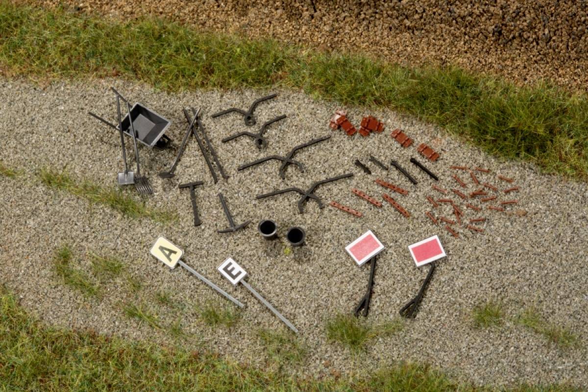 Model railway track accessories on a gravel path, including signs, cable holders and other construction components, distributed across grassy areas.