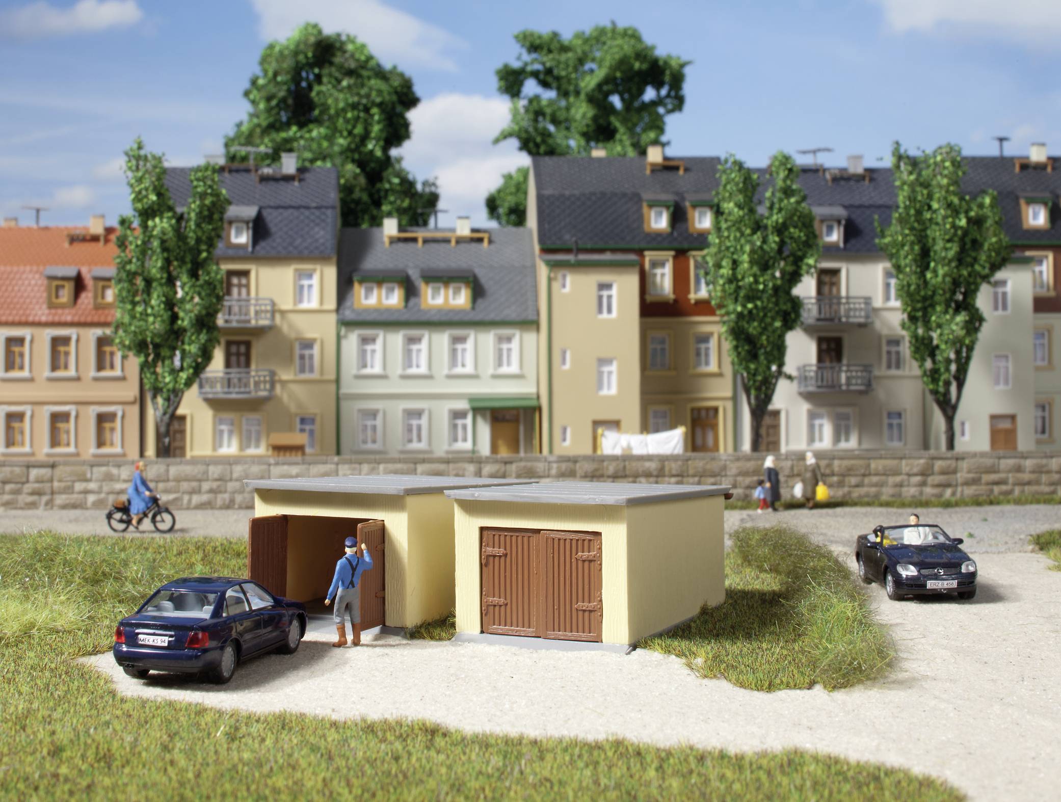 A miniature model of an urban scene with residential houses, trees, cars, and people. One person is standing in front of a garage.