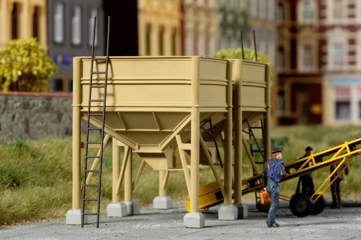 Two grain silos with ladders stand on a yard. A worker wearing a cap walks beside them. Buildings are visible in the background.