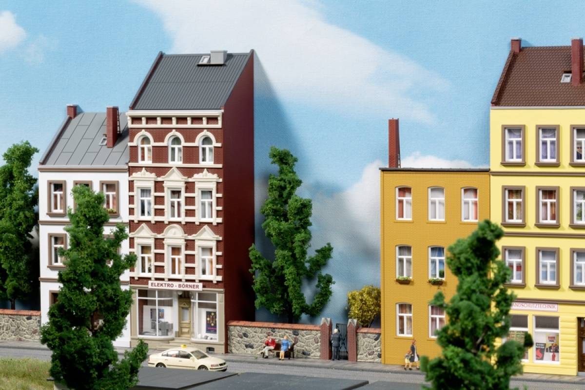Three colourful terraced houses with shops on the ground floor, flanked by trees, against a backdrop of blue sky and clouds.