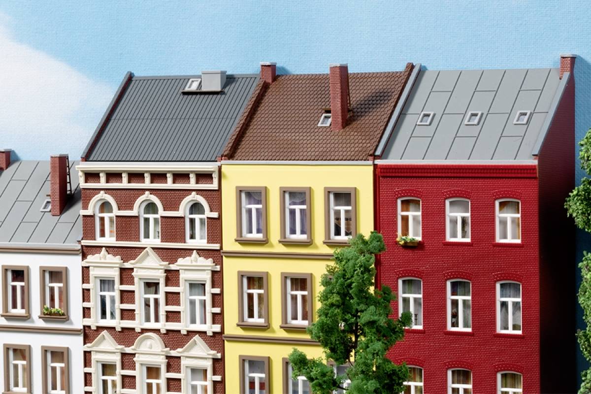 Multicoloured townhouses in a row, each with a unique facade and roof. A tree stands in the foreground. Clear sky.