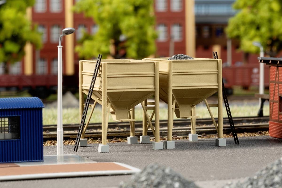 Several large ore bins on a model railway track outdoors, surrounded by rails and buildings on a sunny day.