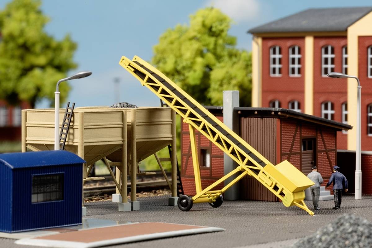 A miniature construction scene with a yellow conveyor belt being observed by two figures. Railway tracks and buildings are visible in the background.
