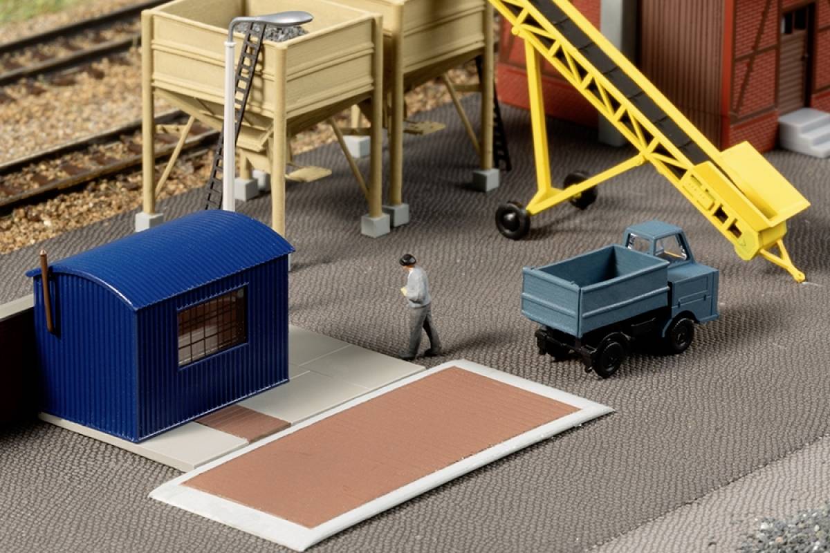 A model lorry next to a small blue building, a figure walking past, with a yellow ramp and railway tracks in the background.
