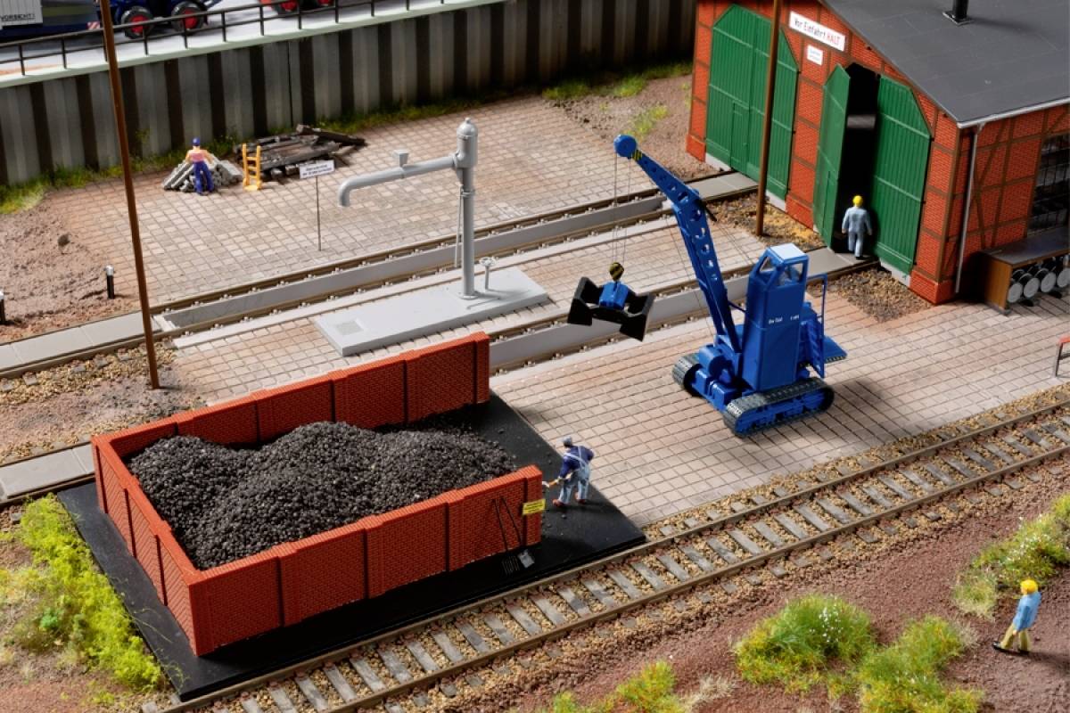 A miniature freight yard shows two workers, a smoking goods wagon loaded with coal, and a blue crane. A building is in the background.