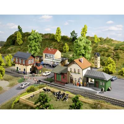 Model railway landscape with a small station, several buildings, cars and cows in a meadow, surrounded by hills and trees.