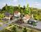 Model railway landscape with a small station, several buildings, cars and cows in a meadow, surrounded by hills and trees.