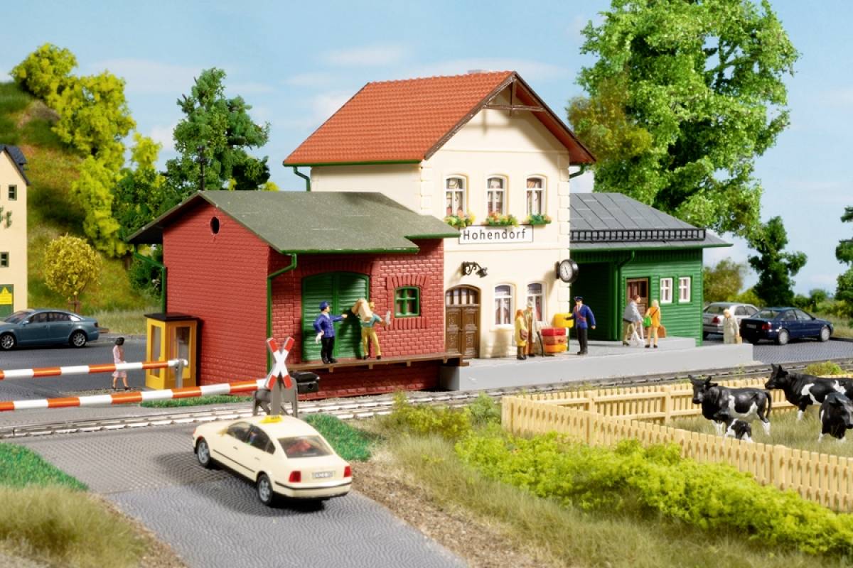 A model railway station with the sign 'Hohendorf'. People on the platform. A level crossing barrier and a car in the foreground.