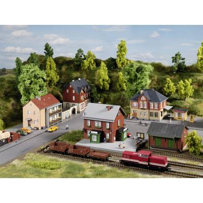 Model railway landscape with a small village, red locomotive and rail freight wagons in the foreground, surrounded by trees and buildings.