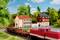 A red model locomotive travels past a small landscape, with two houses and green vegetation in the background.