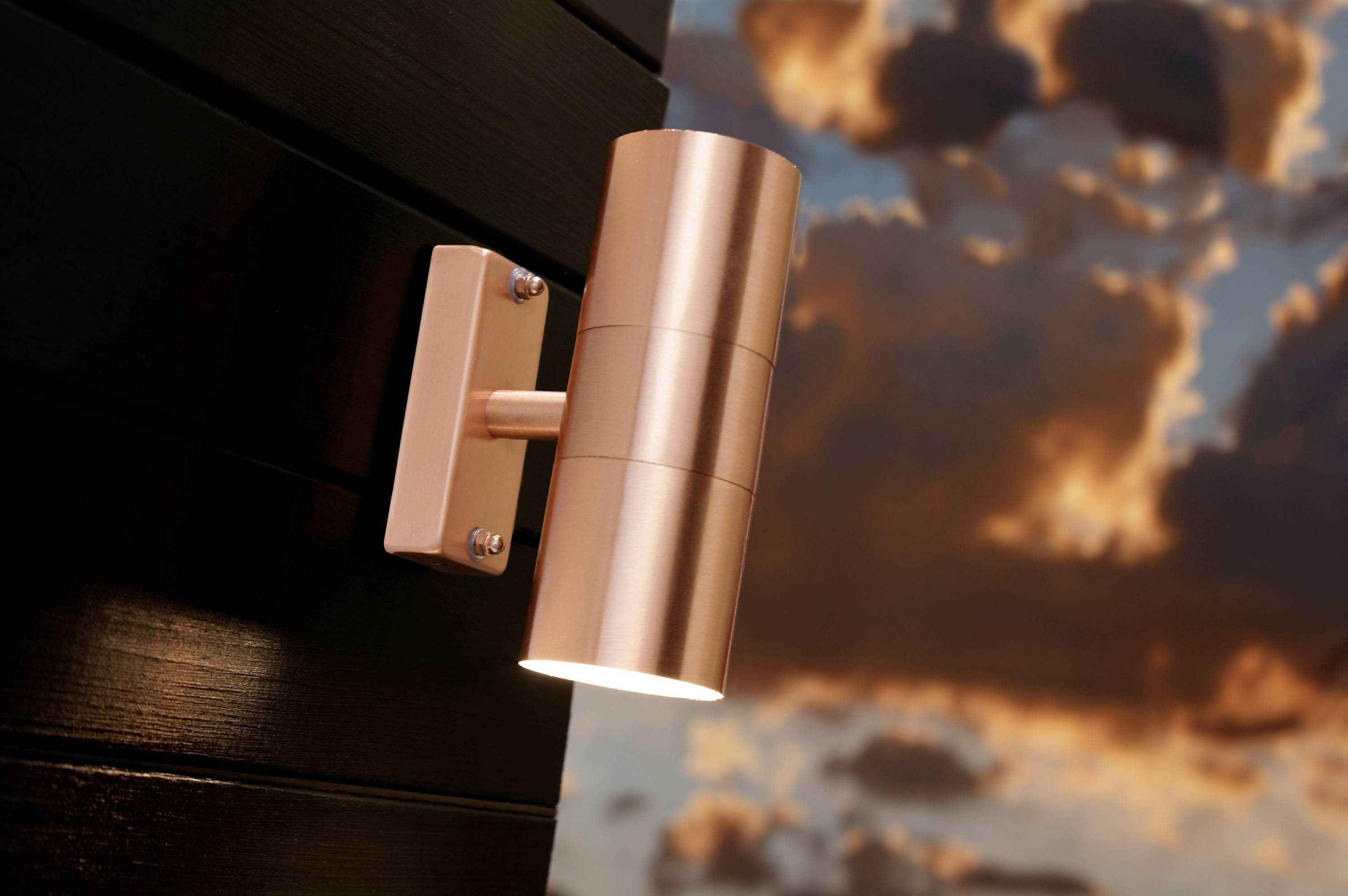 Copper-coloured wall light on a black wall, illuminated at sunset, with clouds in the sky in the background.