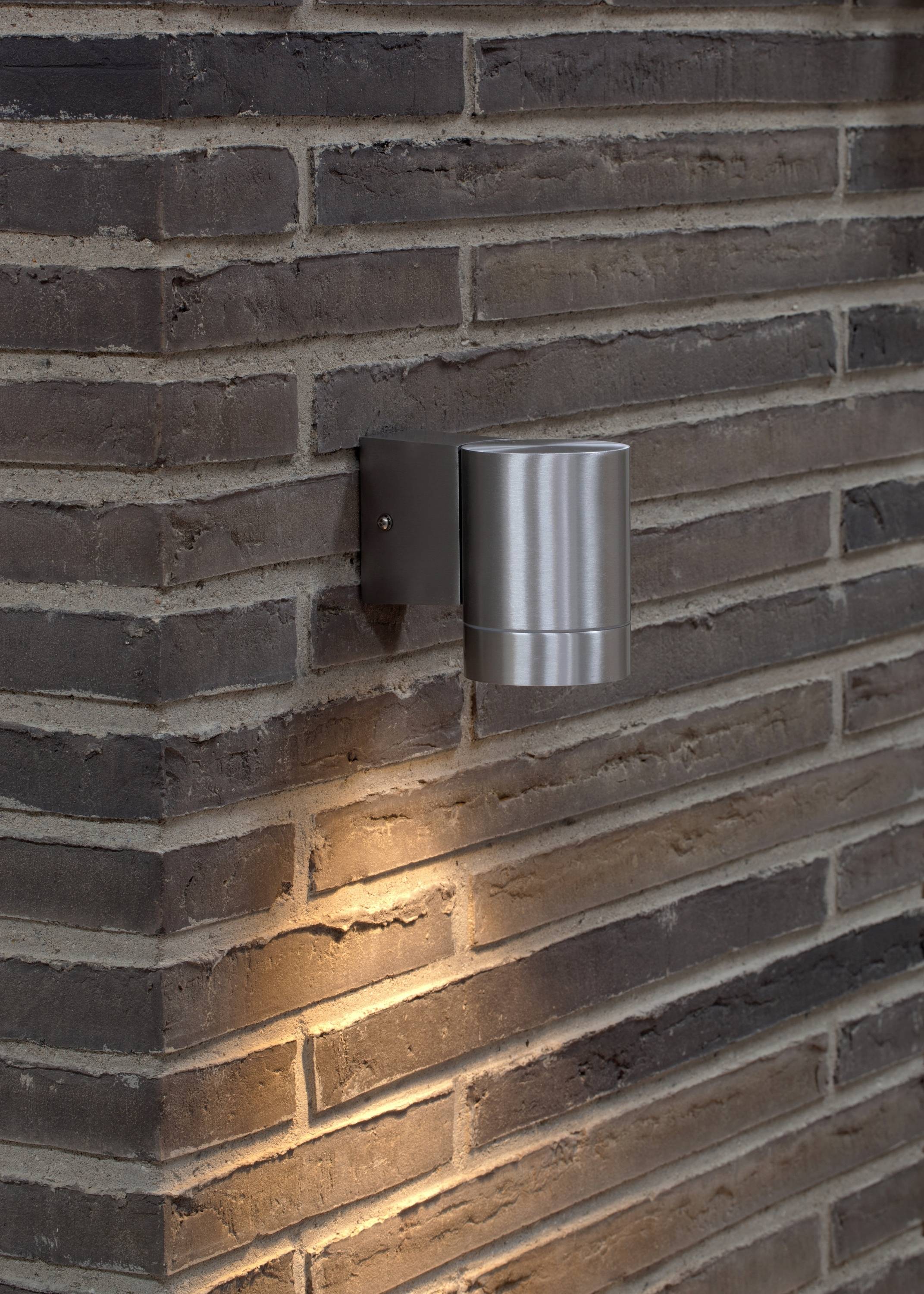 The metal wall light gently illuminates against a brick wall, casting a warm light pattern downwards.