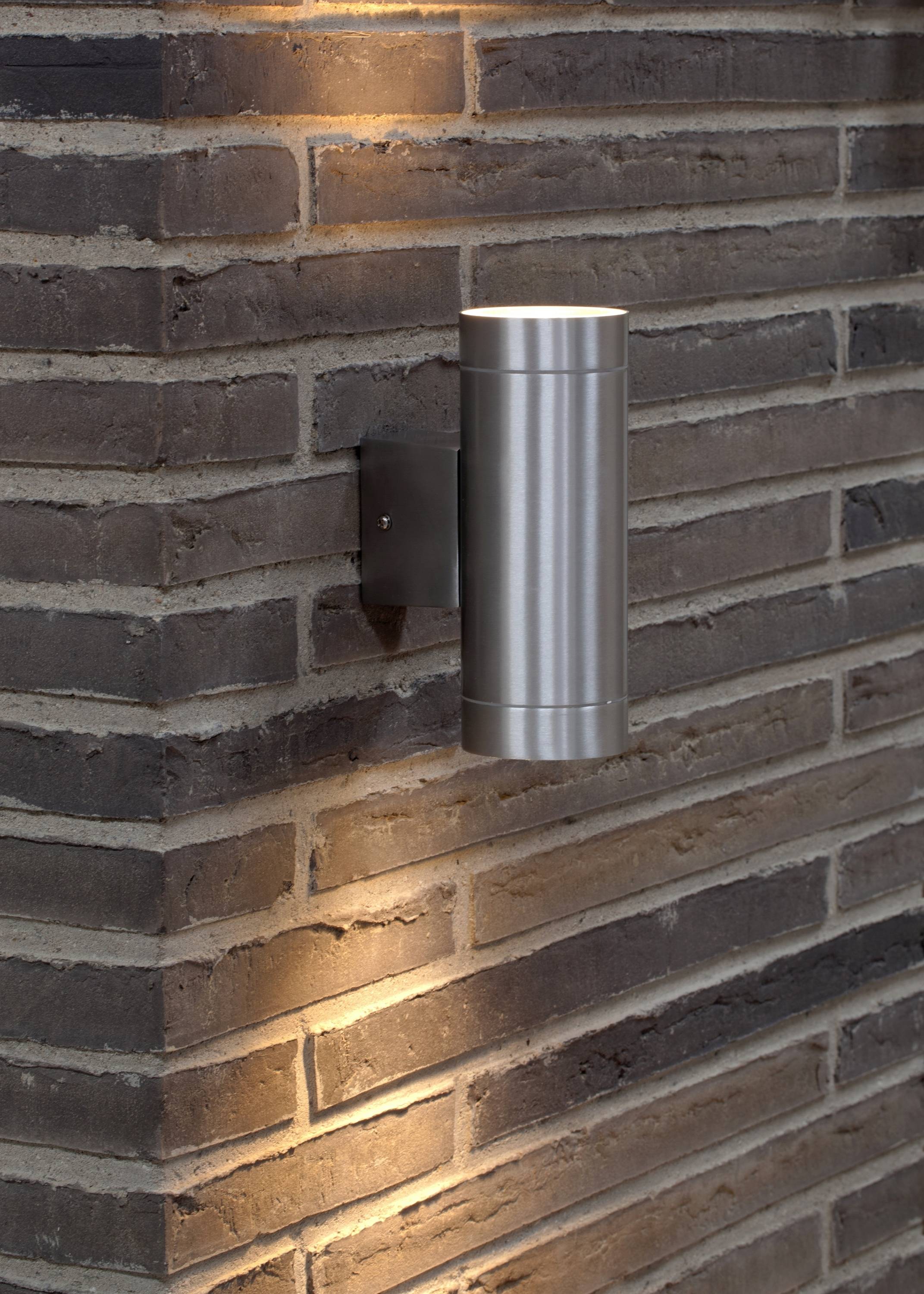 Silver wall light mounted on a brick wall. It emits light upwards and downwards, creating decorative light accents.
