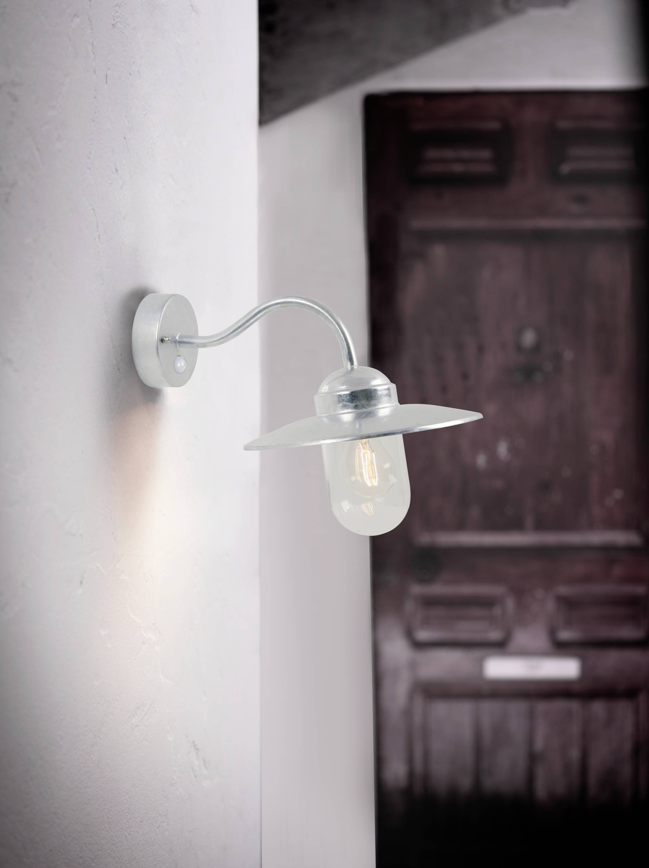 Wall light with a simple metal arm and transparent glass shade, diffused lighting, in front of a blurred wooden door in the background.