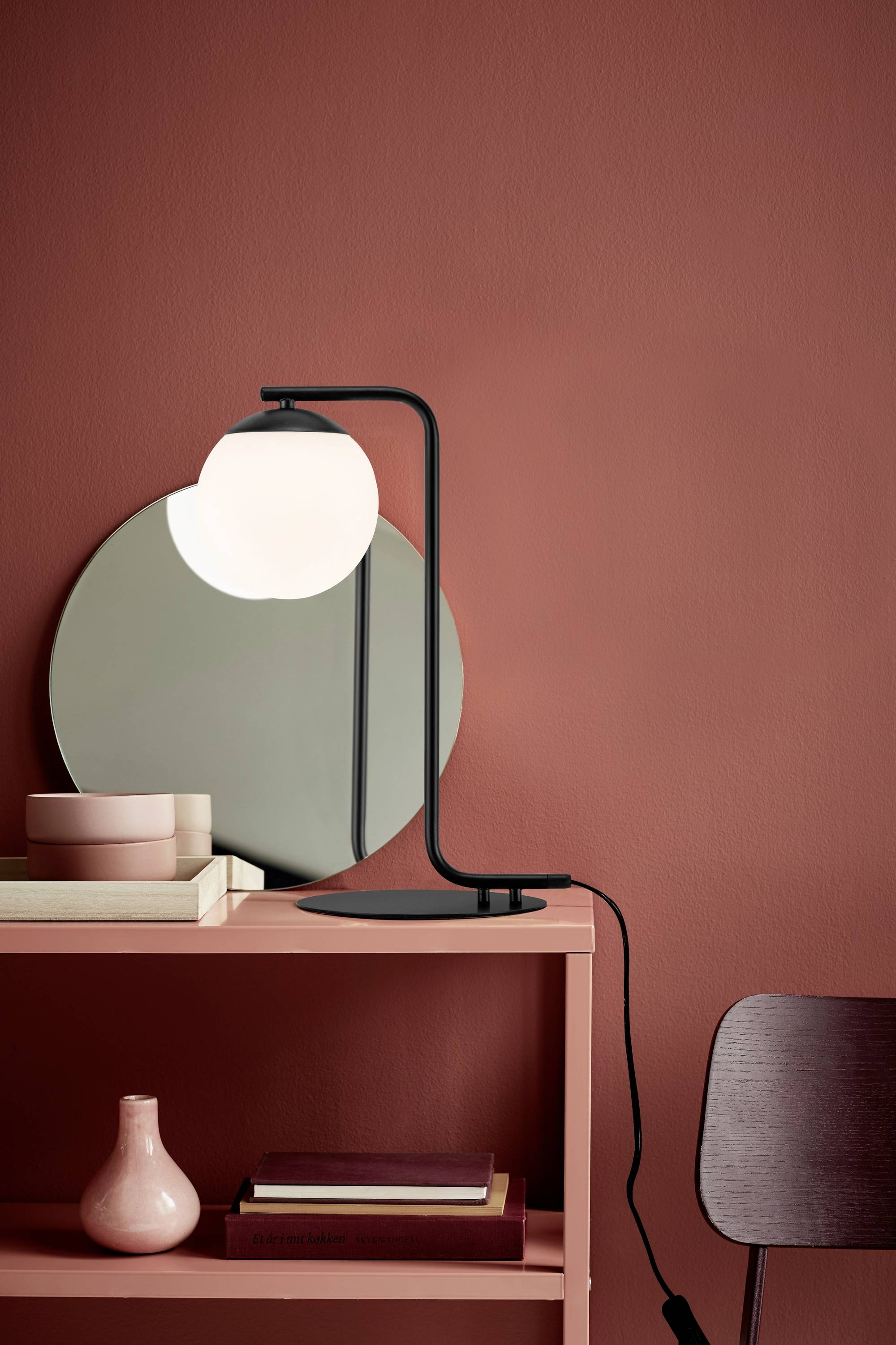 A contemporary table lamp with a round, white shade on a red table. Beside it, a round mirror and books. Background in red.