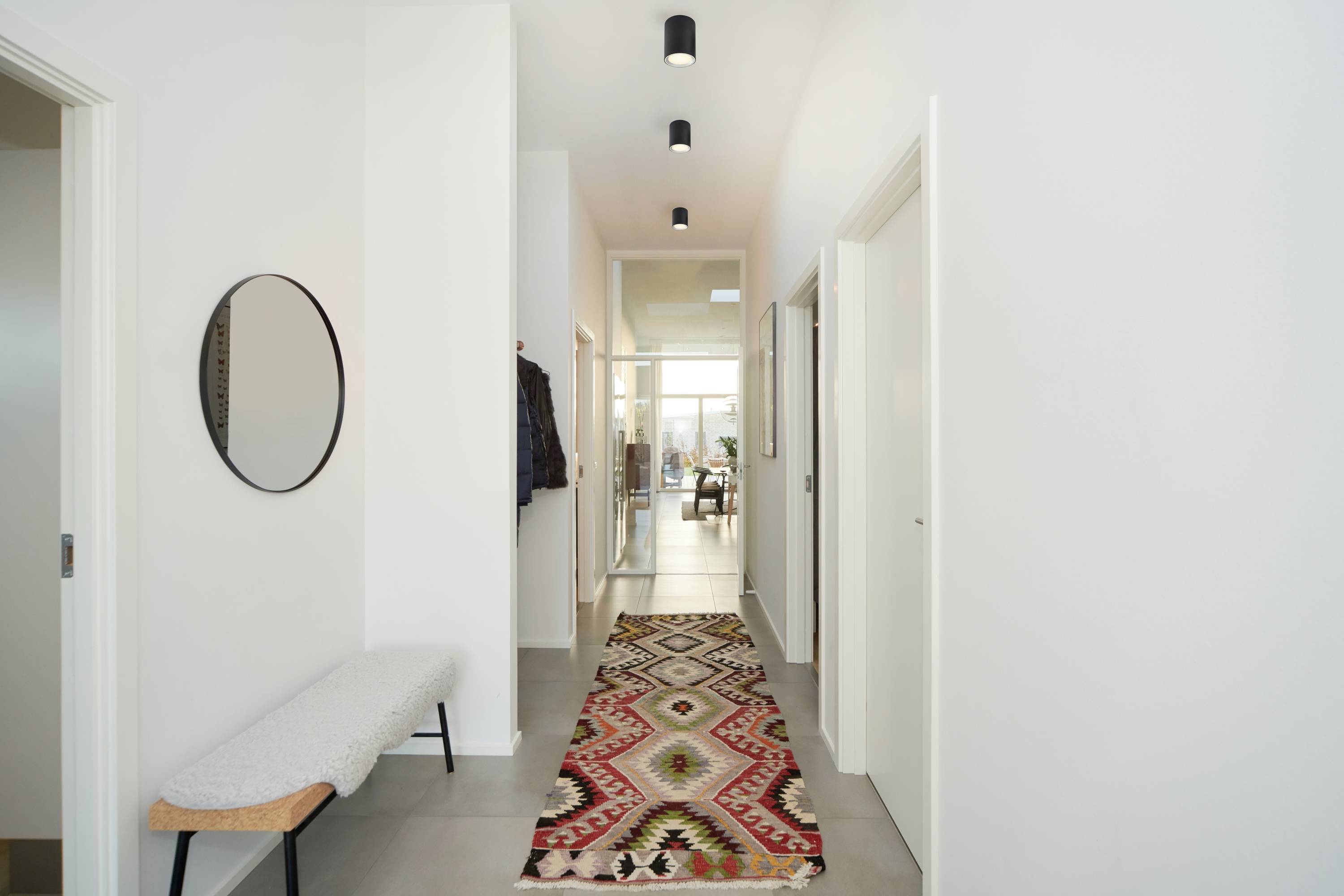 A bright hallway with white-painted walls, a colourful carpet on the floor, a bench on the left, and mirrors on the wall.