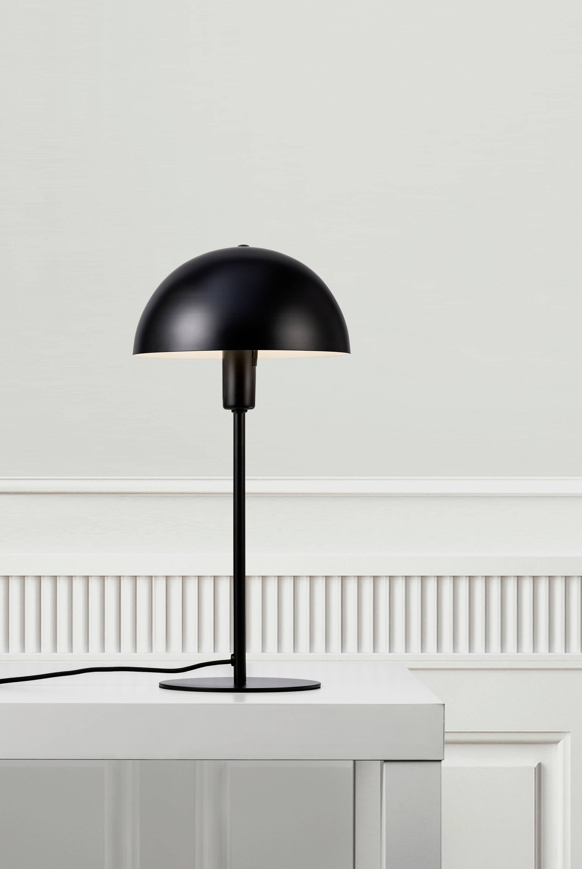 A black table lamp with a semi-circular lampshade sits on a white chest of drawers in front of a light grey wall.