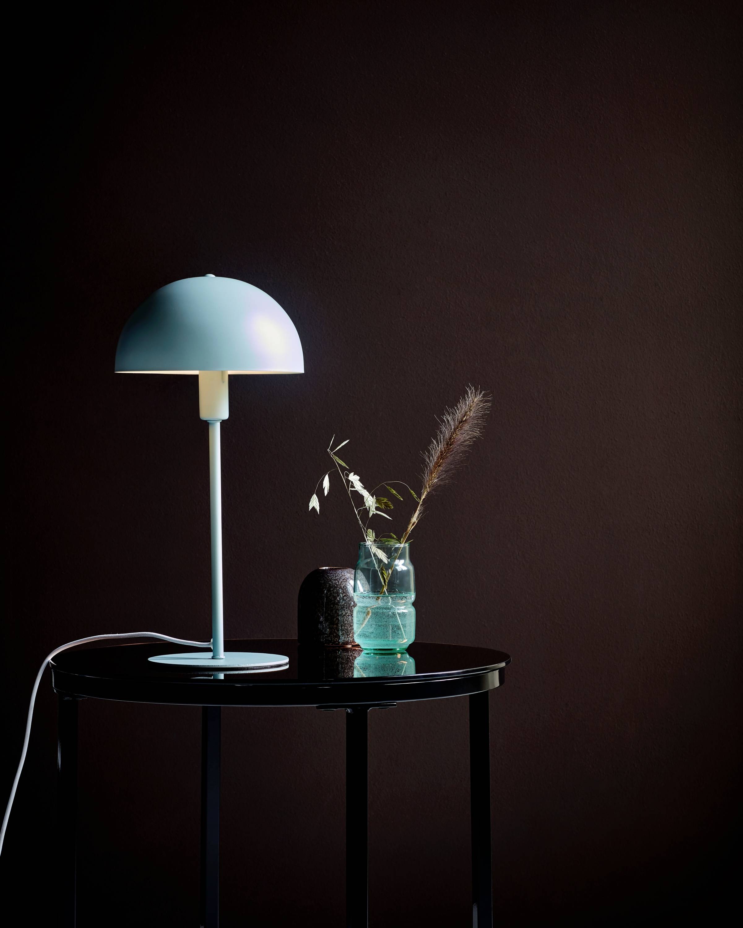 On a small table sits a blue lamp, next to a glass vessel containing dried plants. The background is dark.